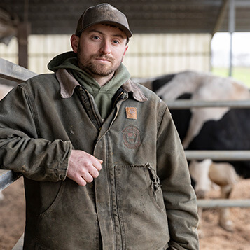 A young male farmer poses for a photo in front of a cow.