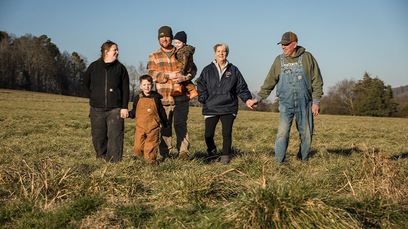 The Barham Jersey Farm family holds hands while posing for a photo in a field of grass.